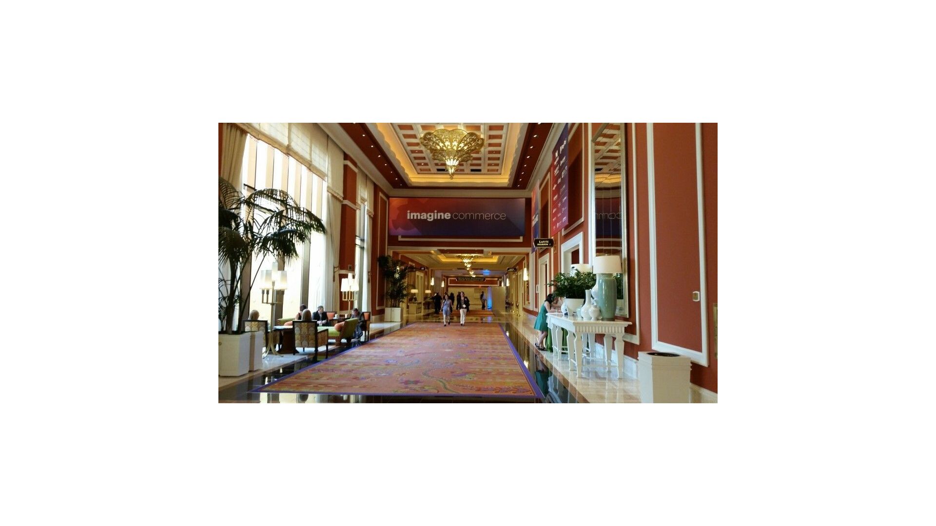 Main hallway of The Wynn Hotel in Las Vegas, Nevada. A chandelier hangs from a high ceiling in an ornately decorated hallway. Over the entry way hangs a sign that reads Imagine Commerce. 