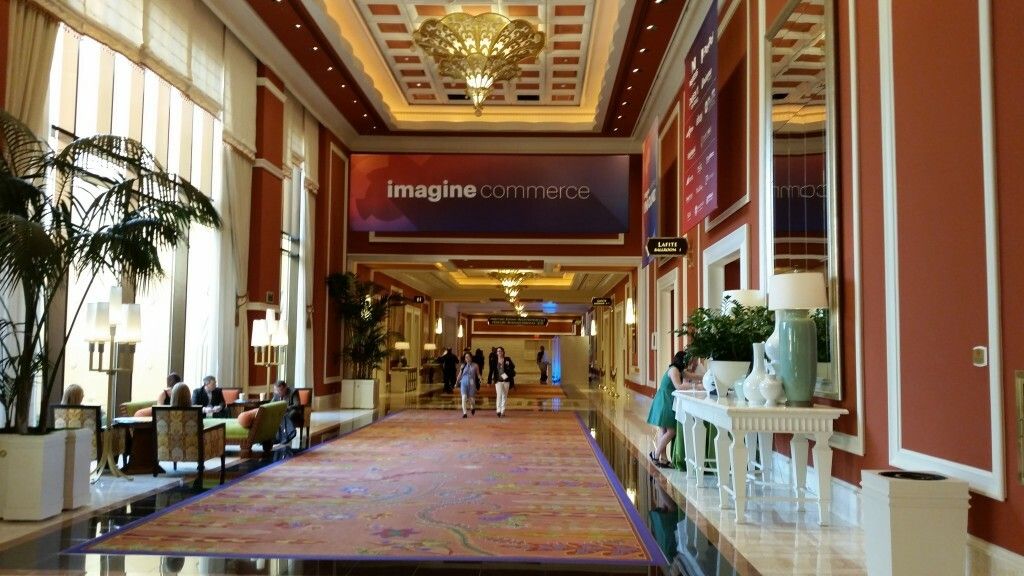 Main hallway of The Wynn Hotel in Las Vegas, Nevada. A chandelier hangs from a high ceiling in an ornately decorated hallway. Over the entry way hangs a sign that reads Imagine Commerce. 