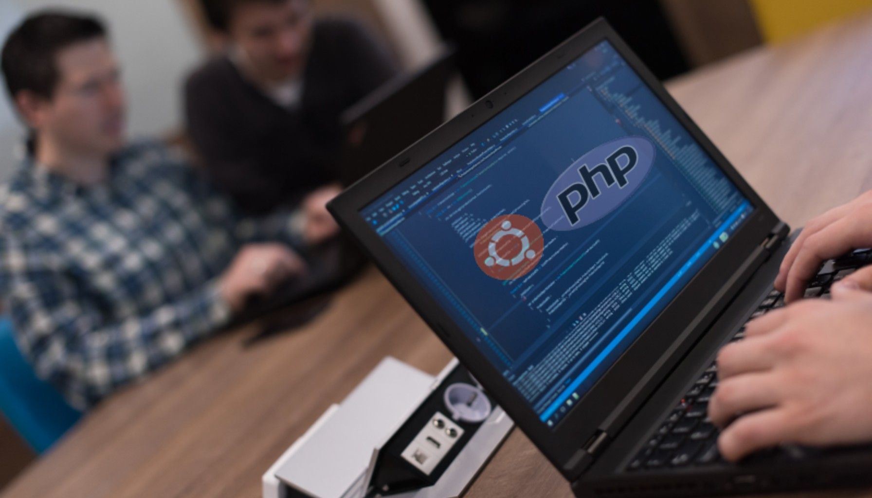 Image of a conference room with people working. A laptop screen is shown with the symbols for Ubuntu and PHP on the screen.